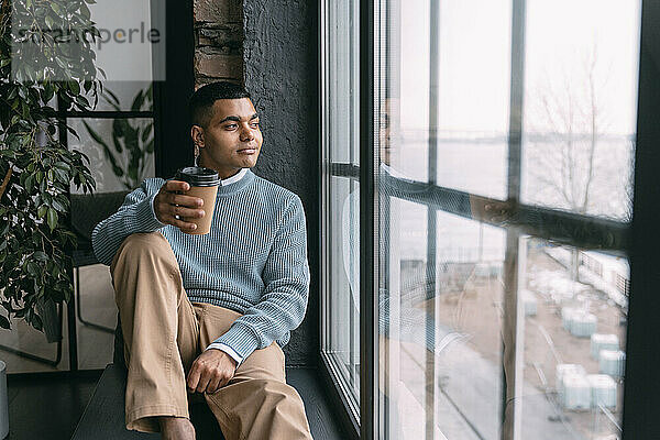 Junger Mann mit Einwegkaffeetasse schaut zu Hause durch das Fenster