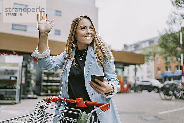 Lächelnde Frau mit Smartphone winkt jemandem zu  der im Laden steht