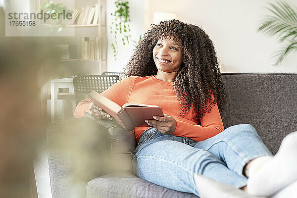 Glückliche Frau mit Buch sitzt zu Hause auf dem Sofa