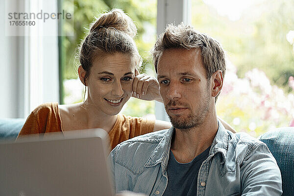 Lächelnde Frau sitzt neben ihrem Freund und benutzt zu Hause Laptop