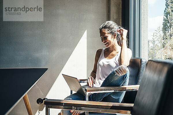 Lächelnde Frau  die zu Hause auf einem Stuhl sitzt und einen Laptop benutzt