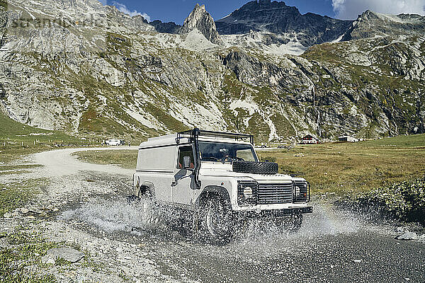 Frau mit Geländewagen fährt an sonnigem Tag durch das Wasser an den Bergen vorbei