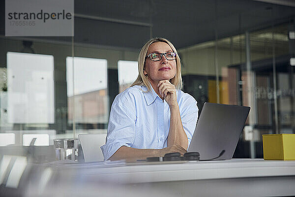 Lächelnde Geschäftsfrau mit Brille und Laptop am Schreibtisch im Büro