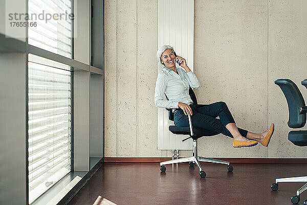 Fröhliche Geschäftsfrau  die am Arbeitsplatz auf einem Stuhl sitzt und mit dem Mobiltelefon spricht
