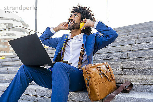 Glücklicher Geschäftsmann  der an einem sonnigen Tag am Laptop sitzt und Musik über Kopfhörer hört