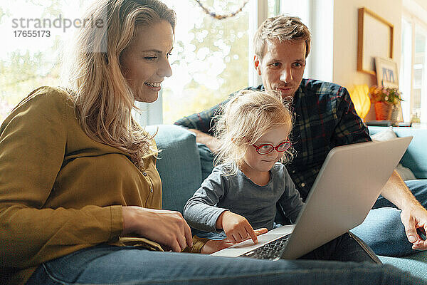 Kleines Mädchen benutzt Laptop und sitzt mit den Eltern zu Hause auf dem Sofa