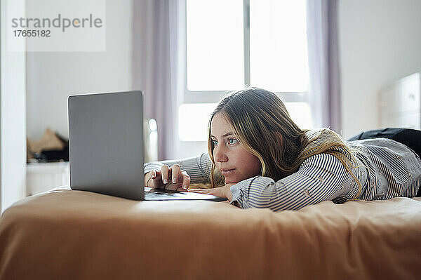 Junge Frau benutzt Laptop und liegt zu Hause im Bett