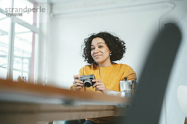Lächelnde Frau mit Kamera sitzt am Tisch im Wohnzimmer
