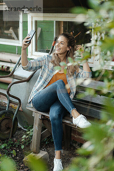 Glückliche Frau macht ein Selfie mit ihrem Smartphone und sitzt auf einer Bank im Hinterhof