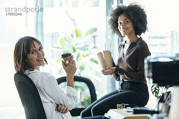 Lächelnde Geschäftsfrau hält Einwegbecher in der Hand und sitzt neben einem Kollegen mit Essen zum Mitnehmen auf dem Schreibtisch im Büro