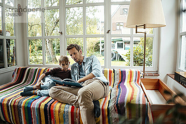 Vater liest seinem Sohn ein Buch vor  der zu Hause auf dem Sofa im Wohnzimmer sitzt