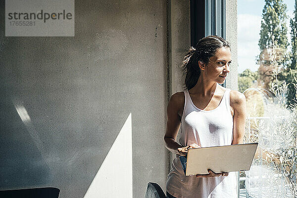 Frau mit Laptop schaut durchs Fenster und steht zu Hause