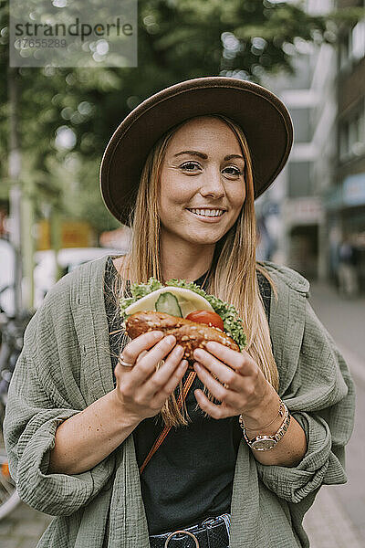 Lächelnde junge Frau mit Sandwich  die auf der Straße steht