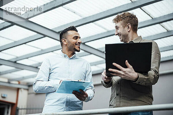 Lächelnder Geschäftsmann hält Laptop in der Hand und diskutiert mit Kollegen am Arbeitsplatz