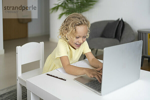 Fröhliches blondes Mädchen sitzt am Tisch im Wohnzimmer und benutzt einen Laptop