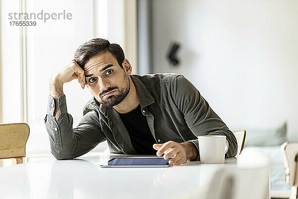 Besorgter Mann mit Tablet-PC lehnt zu Hause am Tisch