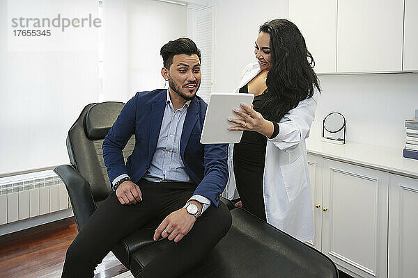 Lächelnder Arzt diskutiert über digitales Tablet mit Patient in ästhetischer Klinik
