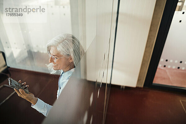 Glückliche Geschäftsfrau benutzt Smartphone im Büro  gesehen durch Glas