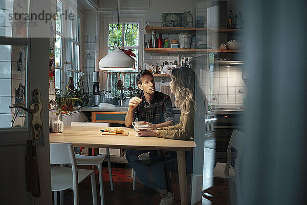 Mann unterhält sich mit Freundin  die am Esstisch in der Küche sitzt
