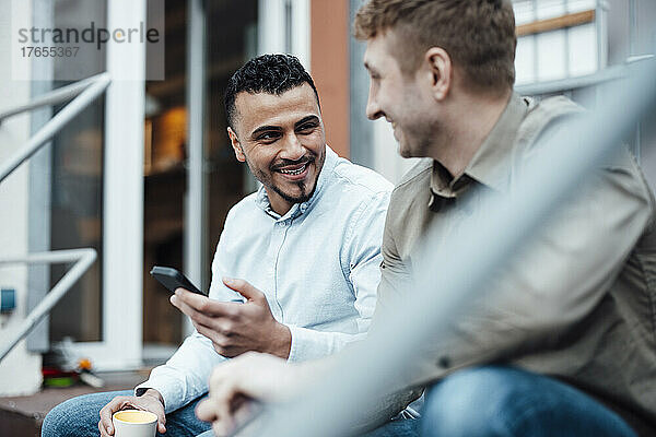 Lächelnder Geschäftsmann hält Mobiltelefon in der Hand und diskutiert mit Kollegen am Arbeitsplatz