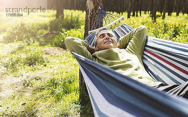 Smiling mature man with hands behind head lying in hammock in forest on sunny day