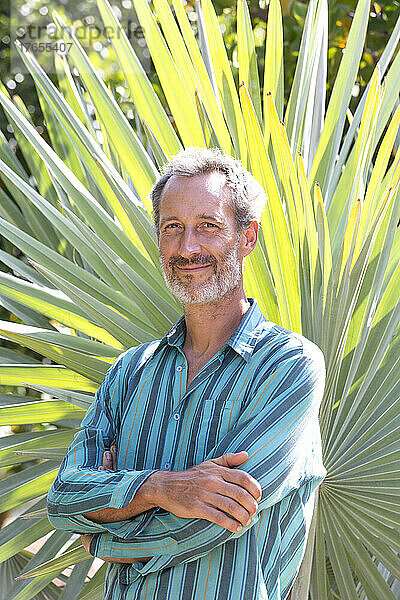 Lächelnder Mann mit verschränkten Armen  der an einem sonnigen Tag vor einem Palmblatt steht