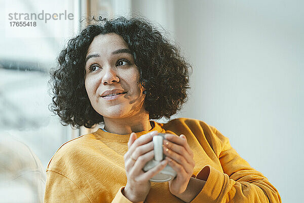 Nachdenkliche Frau mit lockigem Haar hält Kaffeetasse zu Hause