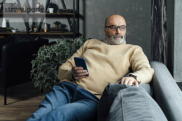 Nachdenklicher älterer Mann mit Mobiltelefon sitzt zu Hause auf dem Sofa