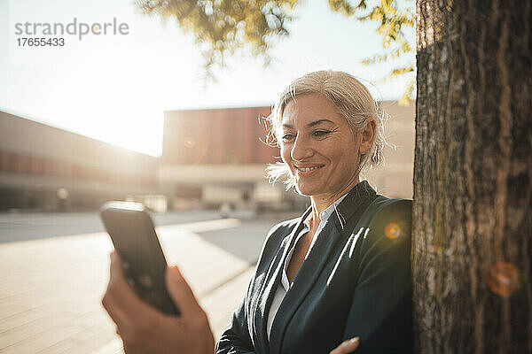 Lächelnde Geschäftsfrau benutzt Smartphone an einem sonnigen Tag am Baumstamm im Büropark