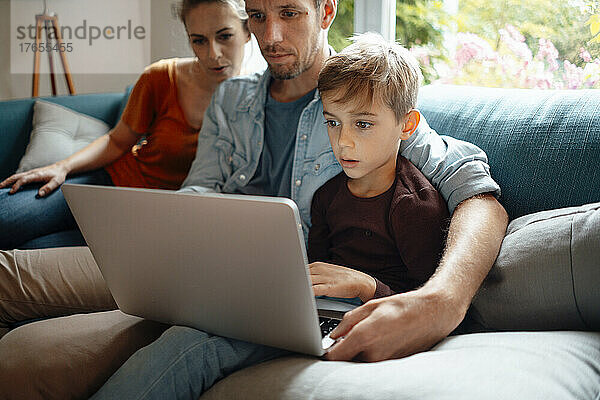 Vater und Sohn teilen sich Laptop und sitzen zu Hause neben einer Frau auf dem Sofa
