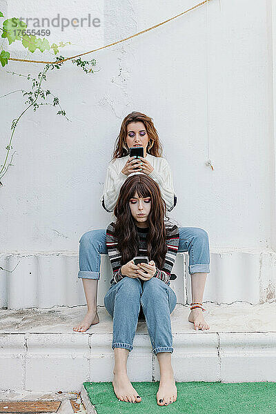 Freunde mit Mobiltelefonen sitzen vor einer weißen Wand im Hinterhof