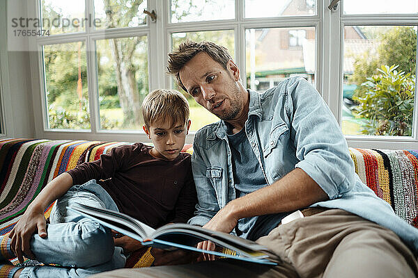 Mann liest seinem Sohn ein Buch vor  der zu Hause auf dem Sofa sitzt