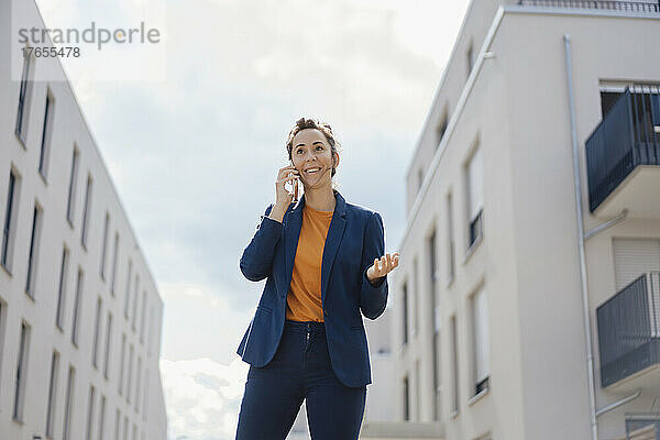 Lächelnde Geschäftsfrau  die an einem sonnigen Tag vor Gebäuden mit dem Handy telefoniert