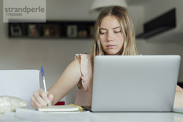 Junge Frau schreibt zu Hause mit Laptop auf Notizblock