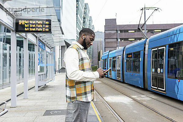 Mann mit kabellosen In-Ear-Kopfhörern benutzt Mobiltelefon an Straßenbahnhaltestelle