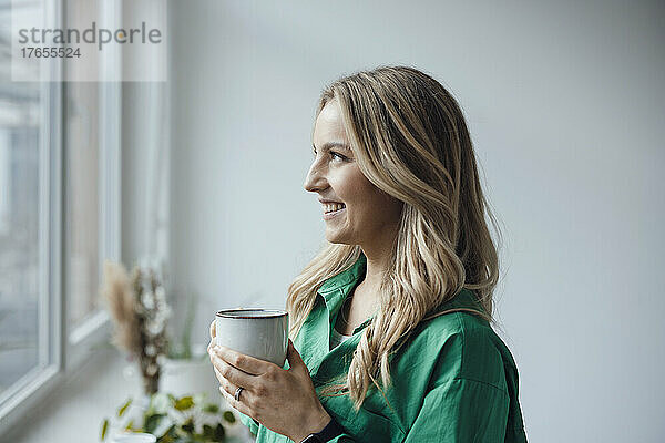 Smiling woman holding coffee cup looking out through window at home