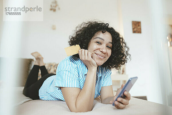 Lächelnde Frau mit Kreditkarte und Smartphone zu Hause im Bett