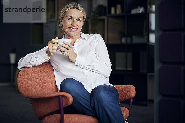 Lächelnde Geschäftsfrau mit Kaffeetasse sitzt auf einem Stuhl im Büro