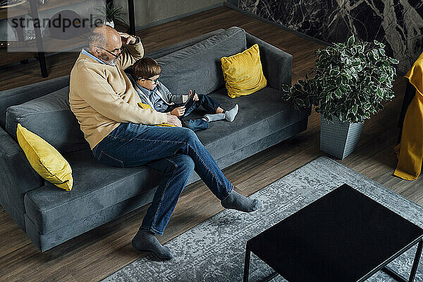 Älterer Mann blickt Enkel beim Lernen über Tablet-PC auf Sofa im Wohnzimmer an