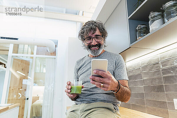 Mann sitzt mit Smoothie und Smartphone auf der Küchentheke in der heimischen Küche