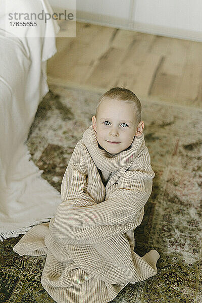 Lächelnder Junge mit übergroßem Pullover sitzt zu Hause