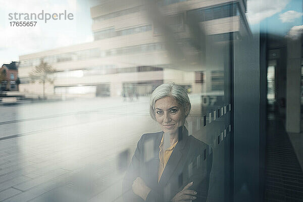 Lächelnde Geschäftsfrau mit verschränkten Armen  gesehen durch Glas  die im Büropark steht