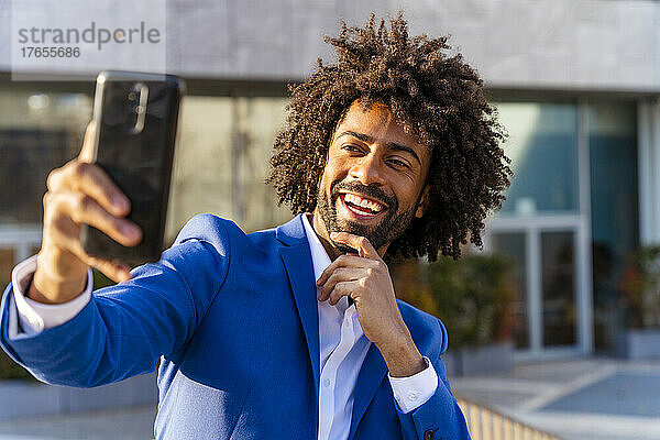 Glücklicher Geschäftsmann mit der Hand am Kinn  der vor dem Gebäude ein Selfie mit dem Mobiltelefon macht