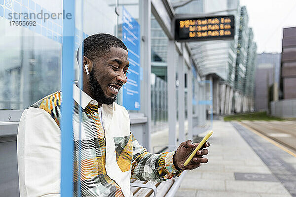 Glücklicher Mann  der über sein Mobiltelefon im Internet surft und an der Straßenbahnhaltestelle sitzt