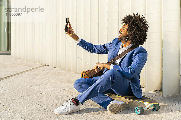 Lächelnder Geschäftsmann macht ein Selfie mit seinem Smartphone und sitzt auf einem Skateboard vor der Wand