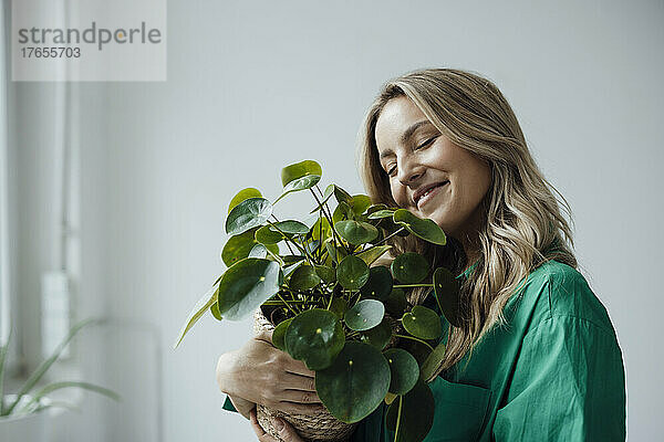 Lächelnde Frau mit geschlossenen Augen umarmt die Pflanze zu Hause