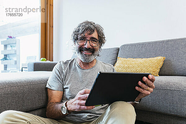 Lächelnder älterer Mann mit Tablet-PC sitzt zu Hause am Sofa