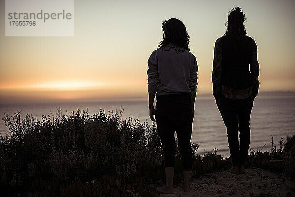 Silhouette zweier Frauen auf einer Meeresklippe  die den Sonnenuntergang in Portugal beobachten