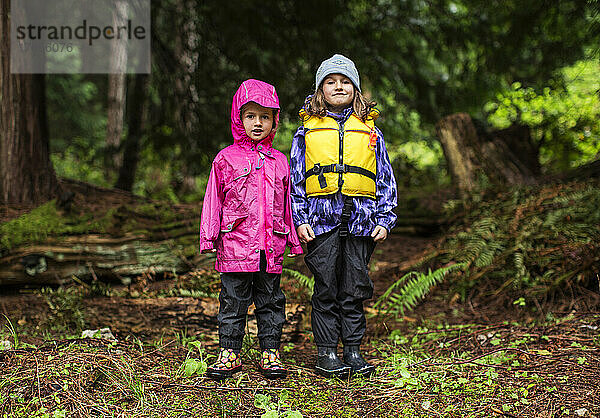 Zwei junge Mädchen in Regenkleidung stehen in einem Wald