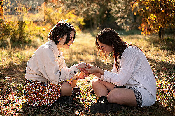 Freundinnen halten Händchen  während sie im Herbst auf Gras im Park sitzen